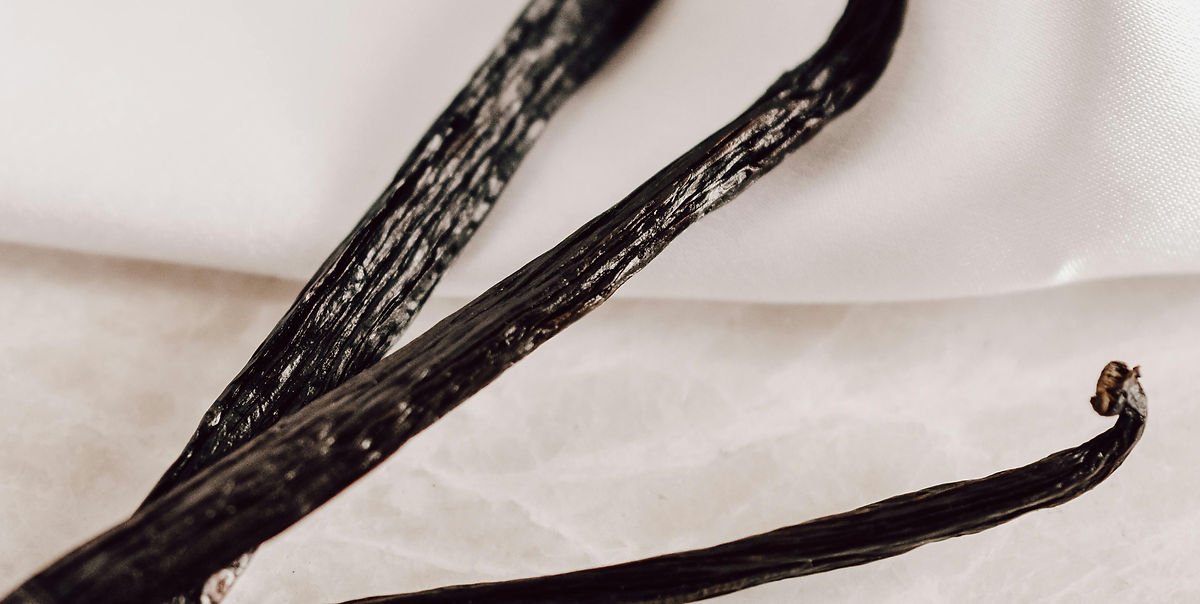 Close-up of dark vanilla pods on a white textured surface. The background is softly lit, with a hint of satin fabric visible.